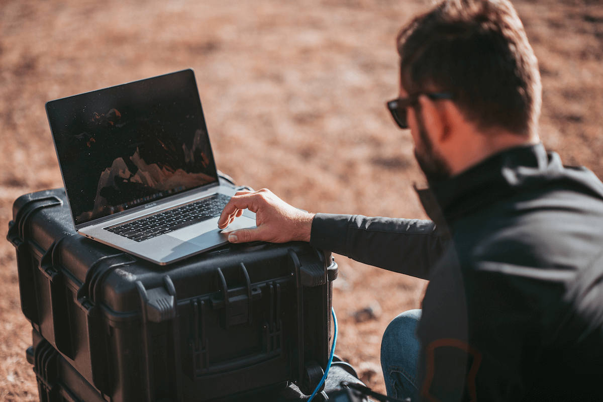 A man sitting in front of an explorer suitcase, engaged with his laptop in a tranquil outdoor scene.